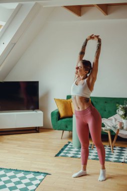 Spor kıyafetleri içinde bir kadın, oturma odasında, parke döşemeli kollarını açıyor.,