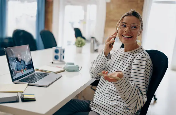 Young smiling female entrepreneur talking by phone with client while sitting in modern office