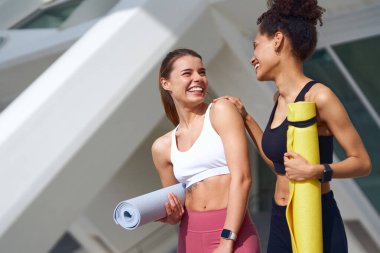 İki kadın dışarıda yoga minderlerini tutarken sohbet ediyorlar.