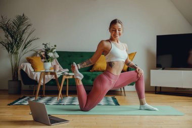 Bir kadın dizüstü bilgisayarın önündeki yoga minderinin üzerinde bacaklarını açıyor ve esneme egzersizleri yapıyor.