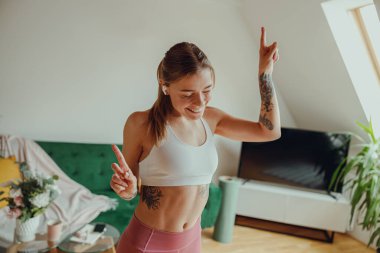 Bir kadın oturma odasında dans ediyor, zarif hareketlerle kollarını havaya kaldırıyor. 