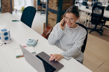 Müşteri ile telefonda konuşan serbest çalışan genç bir bayan bilgisayar üzerinde çalışıyor.