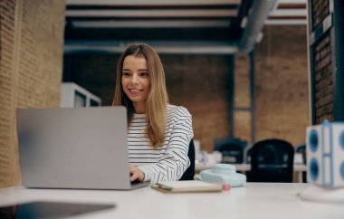 Masada şık bir şekilde otururken dizüstü bilgisayarda çalışan gülümseyen genç bayan yönetici.