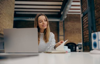 Masada şık bir şekilde otururken dizüstü bilgisayarda çalışan gülümseyen genç bayan yönetici.