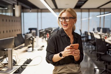 Neşeli bir kadın akıllı telefonunu tutarken etrafındaki hareketli ve dinamik ofis atmosferini gösteriyor.