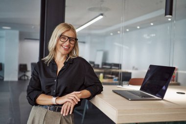 Gözlüklü şık bir kadın, bilgisayarının yanına kurulmuş çağdaş bir ofiste kendine güvenen bir poz veriyor.
