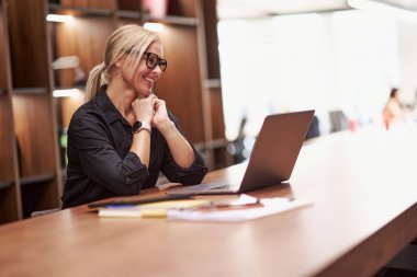 Gülümseyen bir kadın, modern ve çağdaş bir çalışma alanında çalışırken aktif olarak laptopuyla meşgul oluyor.
