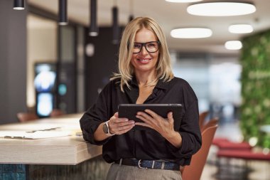 Siyah bluzlu, kendine güvenen bir kadın çağdaş ofis ortamında tabletiyle etkileşime geçiyor..