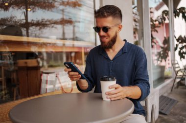 Şık güneş gözlüklü genç sakallı bir adam şık bir kafede kahvenin tadını çıkarırken akıllı telefonuyla çok meşgul.