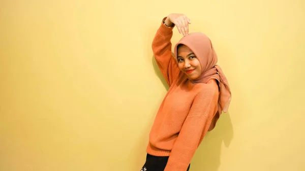 兴奋的亚洲穆斯林妇女穿着橙色毛衣 头戴头巾 指着旁边被橙色背景隔开的抄袭空间 — 图库照片