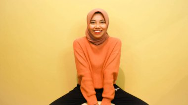 Çekici, güzel Asyalı kadın, turuncu arka planda izole edilmiş mutluluk hissiyle sandalyede oturuyor..