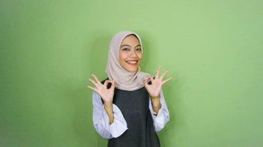 Kendinden emin, neşeli, Asyalı genç bir kadın başparmağından memnun, onay ver, iyi seçimi sev, gülümse, mutlu ol, olumlu düşün, yeşil arka planda dur.