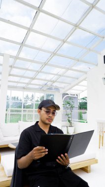 Siyah üniformalı yakışıklı Asyalı adam evde kanepede otururken kitap okuyor.