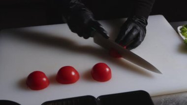 Siyah eldivenli aşçı büyük bir bıçakla domatesi keser.
