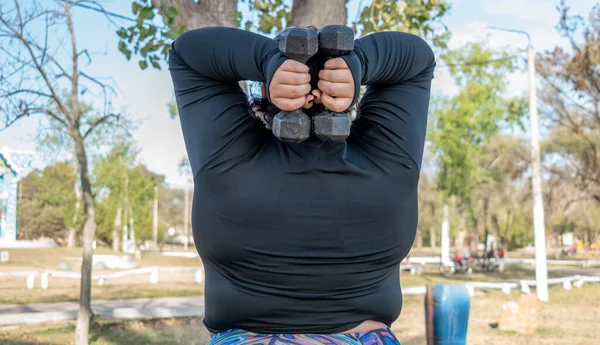 Young fat woman exercising with dumbbells in the park by the lak