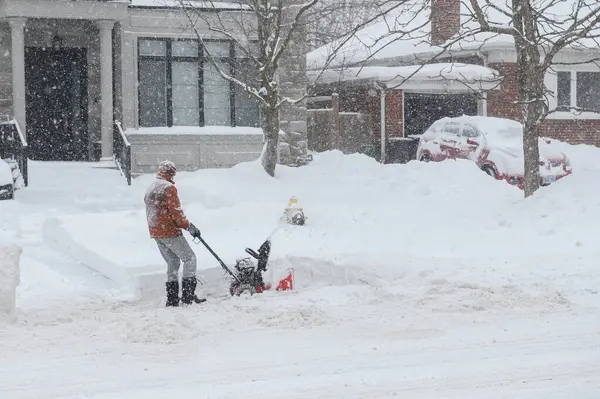 Toronto, ON, Kanada - 27 Şubat 2025: Bir adam varoş mahallesinde yoğun kar yağışı sırasında garaj yolundaki derin karı temizlemek için kar temizleme makinesi kullanıyor.