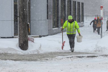 Toronto, ON, Kanada - 27 Şubat 2025: Bir kişi kar fırtınası sırasında karla kaplı ağaçların ve tuğla evlerin yakınındaki bir yolda kar kürüyor.