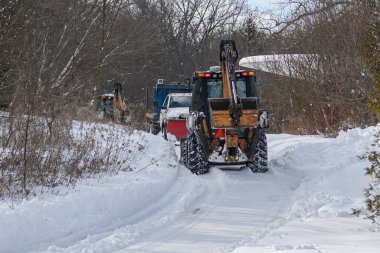 Toronto, ON, Kanada - 27 Şubat 2025: Güvenlik ekipmanlı iki işçiyle kar küreme aracı kar kaplı kırsal bir yolu temizliyor, soğuk bir kış gününde taze karı kenara itiyor.