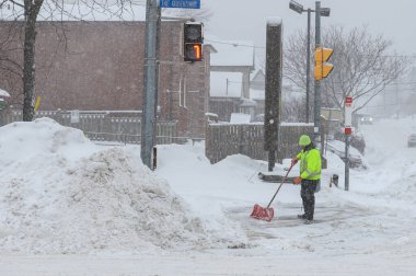 Toronto, ON, Kanada - 27 Şubat 2025: Bir kişi kar fırtınası sırasında karla kaplı ağaçların ve tuğla evlerin yakınındaki bir yolda kar kürüyor.