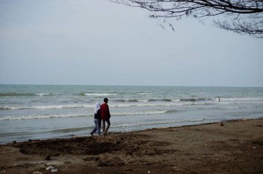 Daytime view of beach with local people , pantai utara jawa, di kota Pemalang, ombaknya kecil, nampak pasangan couple berjalan di pinggir pantai, sedang berlibur, di pantai Widuri, Pemalang, Jawa Tengah, Indonesia, pada siang hari 