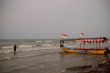 Local man standing beach of wavy sea at daytime, perahu nelayan di pinggir pantai Widuri Pemalang, Indonesia, digunakan untuk angkutan wisata laut, wisatawan diajak naik perahu ke tengah laut, ombaknya kecil, resiko kecil membayar sejumlah uang 