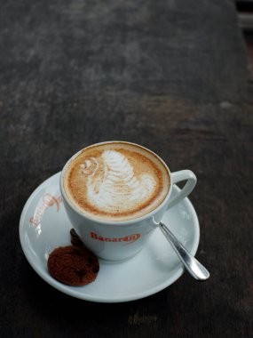 Caffe Latte. Cup of coffee with a sweet cookie. capucino kopi dari Resto Banaran 9 kopi di Semarang. nampak disajikan dengan wadah cangkir berwarna putih , lambar putih, ditambahkan biskuit coklat dan biji kopi, juga disiapkan sendok untuk mengaduk.