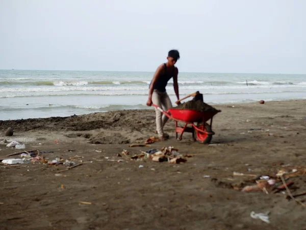 Young asian man with wheelbarrow cleaning sandy beach at daytime , pantai utara jawa, di kota Pemalang, ombaknya kecil, nampak pekerja di pinggir pantai, sedang mencari pasir, di pantai Widuri, Pemalang, Jawa Tengah, Indonesia, pada siang hari 