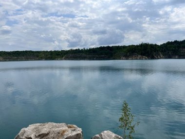 Gökyüzünün yansıması ile sakin mavi göl ve yoğun yeşil ormanlarla çevrili uzak kayalık uçurum