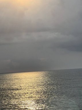 Sakin deniz yüzeyine yansıyan altın güneş ışığı dramatik bulutlu gökyüzünün altında huzurlu okyanus manzarası yaratıyor.