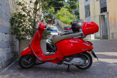 Bellano, LombardyItaly-06.16.2023. Kuzey İtalya 'da Bellano kasabasındaki bir kilisenin önüne park etmiş kırmızı bir Vespa scooter. Güneşli Haziran günü.