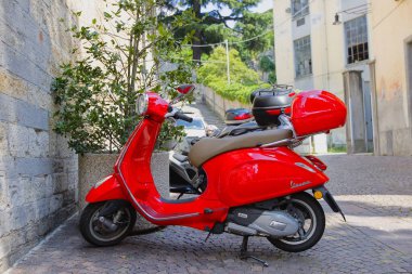 Bellano, LombardyItaly-06.16.2023. Kuzey İtalya 'da Bellano kasabasındaki bir kilisenin önüne park etmiş kırmızı bir Vespa scooter. Güneşli Haziran günü.