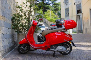 Bellano, LombardyItaly-06.16.2023. Kuzey İtalya 'da Bellano kasabasındaki bir kilisenin önüne park etmiş kırmızı bir Vespa scooter. Güneşli Haziran günü.