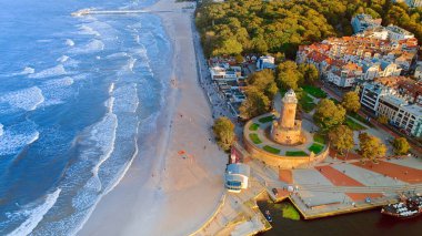 Drone photo captures Kolobrzeg's maritime charm, featuring the iconic lighthouse, cerulean sea, turbulent waves, a distant pier, and autumnal hues on the trees. clipart