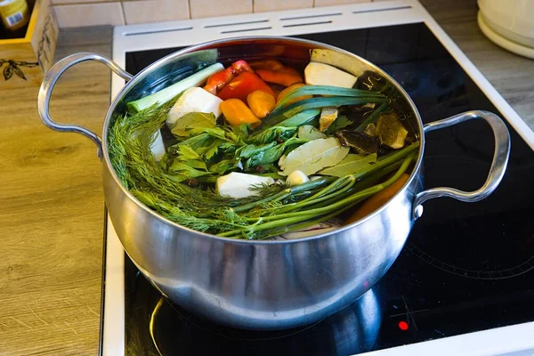 Stainless steel pot on electric stove with ceramic cooktop cooking organic chicken broth. Ingredients: veggies, chicken, bell pepper, celery, leeks, celery leaves, bay leaves, garlic, onion, carrots.