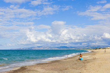 Platamona Sahili, Sardinya, İtalya 'da, bir Sardunyalı balıkçı bir çubukla resmedilir. Görüntü sahili, uzak dağları ve mavi gökyüzündeki çarpıcı bulutları gösteriyor..