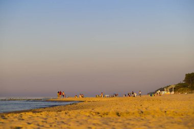 Mrzezyno 'da bir sahil, Batı Pomeranya Voyvoda, Polonya, altın saat içinde ele geçirildi, sıcak güneş ışığıyla güzelce aydınlatıldı. Gezen ve güneşlenen insanlar, temiz gökyüzü, kumullardaki bitki örtüsü, sahile çıkan merdivenler..