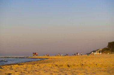 Mrzezyno 'da bir sahil, Batı Pomeranya Voyvoda, Polonya, altın saat içinde ele geçirildi, sıcak güneş ışığıyla güzelce aydınlatıldı. Gezen ve güneşlenen insanlar, temiz gökyüzü, kumullardaki bitki örtüsü, sahile çıkan merdivenler..