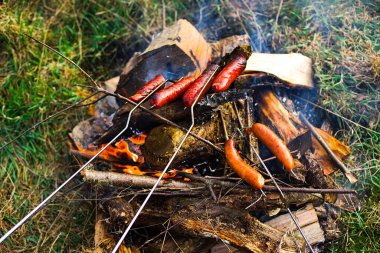 Geleneksel tarzda sosisler odun közleriyle çimenli bir şenlik ateşinde kızarıyor ve beyaz duman yayıyor. Sosislerin güzel bir altın-kahverengi rengi vardır..
