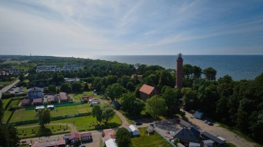 Gski deniz feneri, kumlu Baltık plajı ve çevresindeki köyün insansız hava aracı görüntüsü, hepsi parlak, sıcak bir manzara içinde..
