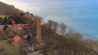 A drone shot displays Gaski beach, West Pomeranian Voivodeship, Poland, with a red brick lighthouse, Baltic Sea, sandy beach, leafless dune trees, holiday cottages, hotels, and homes. Possibly calm sea. Captured in February winter. clipart