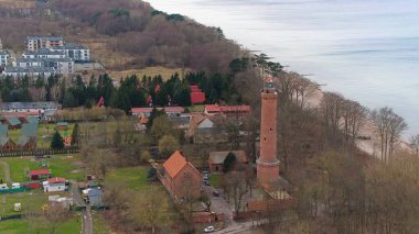 A drone shot displays Gaski beach, West Pomeranian Voivodeship, Poland, with a red brick lighthouse, Baltic Sea, sandy beach, leafless dune trees, holiday cottages, hotels, and homes. Possibly calm sea. Captured in February winter. clipart