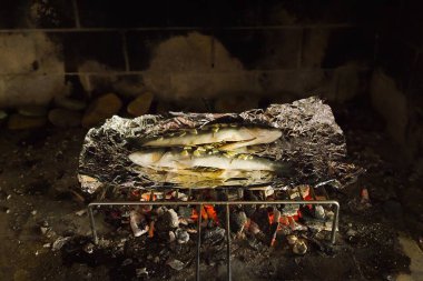 İki levrek, sarımsak dilimleri ve taze biberiye filizleriyle süslenmiş geleneksel İtalyan ızgarasının üzerinde folyonun üzerinde dinleniyor. İçinde tereyağı, sarımsak ve biberiye var. Altında, kızgın kömürler yoğun bir şekilde parlıyor..