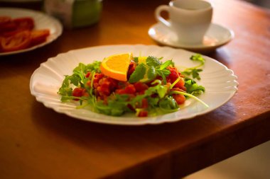 White plate holds tuna tartare with fresh arugula, mint, and citrus slices, set on a wooden table; breakfast scene softly blurred in the background. clipart