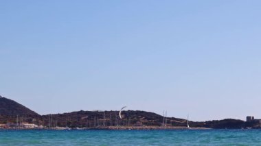 Rüzgarlı bir günde yakalanan bir uçurtma sörfçüsü, Villasimius, Sardinya 'daki Campulongu Körfezi sularında bir marina, yatlar ve Capo Carbonara ile arka planda gezinir..