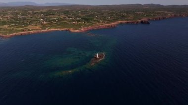 Sardinya 'nın Faro Mangiabarche deniz feneri, engebeli kayalara tünemiş, sakin mavi denizlere ve yumuşak dalgalara bakıyor. İnsansız hava aracı görüntüleri, sıcak ve güneşli gökyüzünün altında güzel bir sahil manzarası gösteriyor..