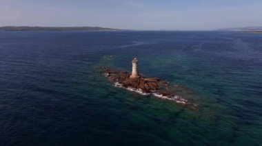 Sardinias Faro Mangiabarche deniz feneri bir sahil mücevheridir. İnsansız hava aracı beyaz kulesini engebeli kayalar, mavi sular ve güneşli bir gökyüzünün altında hafif dalgalar arasında yakalar..