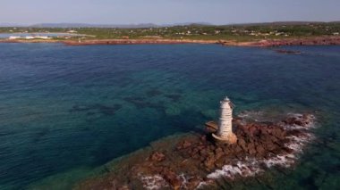 Sardinias Faro Mangiabarche deniz feneri bir sahil mücevheridir. İnsansız hava aracı beyaz kulesini engebeli kayalar, mavi sular ve güneşli bir gökyüzünün altında hafif dalgalar arasında yakalar..