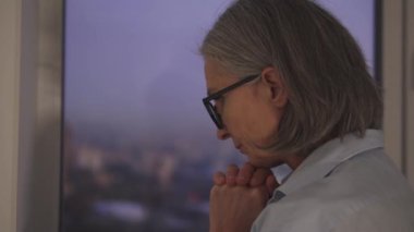 Gri saçlı, olgun bir kadın Tanrı 'yla konuşuyor, kollarını kavuşturup dua ediyor ve dışarı bakıyor.