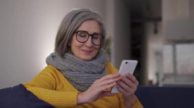 Kendinden emin ve şık olgun bir kadın, evinden akıllı telefon üzerinde çalışarak ek para kazanıyor. Emekli insanlar için kolay gelir, reklam.