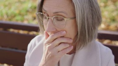 Gözlüklü ve şık montlu endişeli olgun bir kadın ellerini ovuşturuyor, sonbahar parkında bankta oturuyor, zor hayatın çözümünü arıyor, kafa karışıklığını hissediyor.